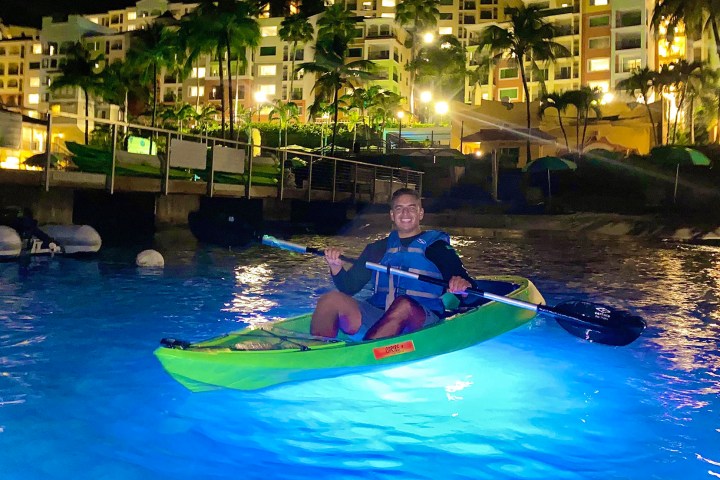 Person kayaking in glowing blue water near lit buildings at night.