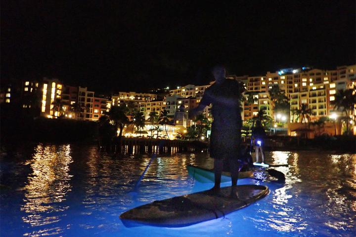 Person paddleboarding on illuminated water at night near lit buildings.