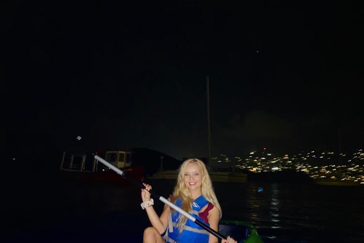 Person kayaking at night on a dark lake, city lights in the background.