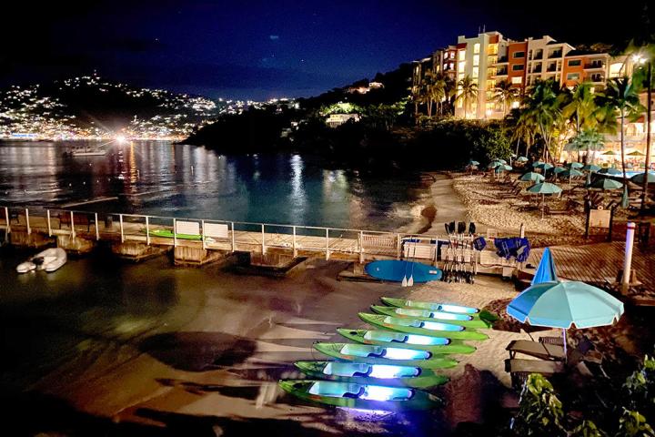 Beach at night with kayaks, umbrellas, and illuminated buildings.