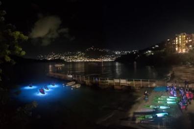 Night view of bay with illuminated water and kayaks, city lights in the background.