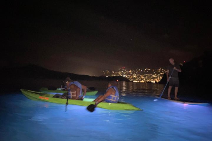 People kayaking and paddleboarding on glowing water at night with city lights in the background.
