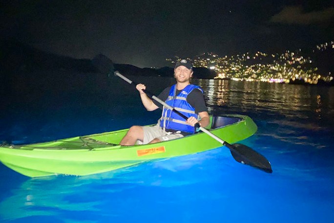 Person kayaking at night over bioluminescent water, city lights in the background.