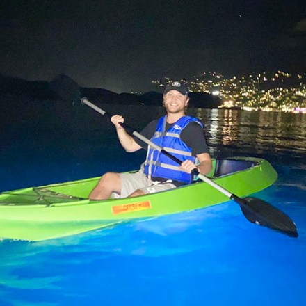 Person kayaking at night over bioluminescent water, city lights in the background.