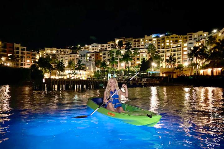 Two people kayaking on glowing water at night with lit buildings in the background.