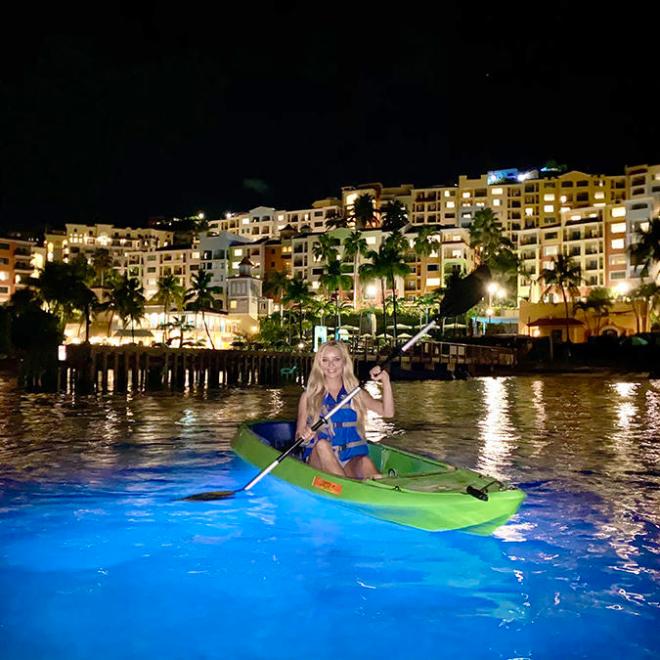 Two people kayaking on glowing water at night with lit buildings in the background.