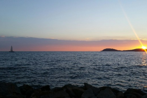 Sunset over ocean with a distant sailboat and rocky shore