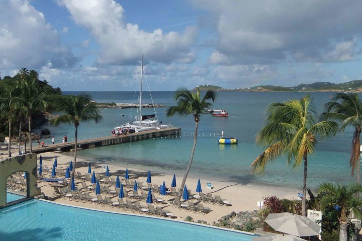 Beachfront with palm trees, sailboat, and lounge chairs under blue umbrellas.
