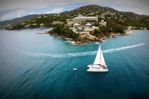 Sailboat near a lush island with buildings and hills in the background.