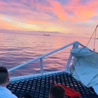 People relaxing on a boat at sunset with pink sky and calm sea.