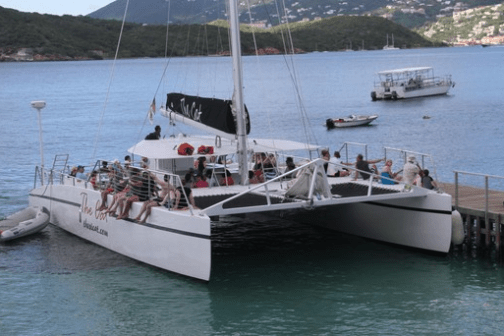 Large catamaran docked with passengers on board, surrounded by calm water and hills in the background.