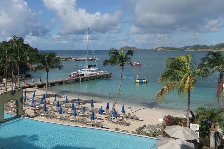 Beach scene with palm trees, loungers, umbrellas, pool, dock, sailboats, and cloudy sky.