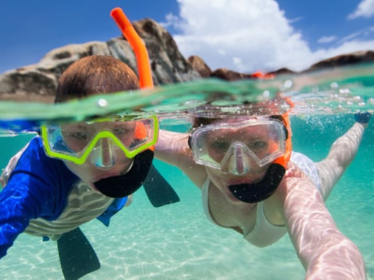 Two people snorkeling underwater with clear sky and rocks in the background.