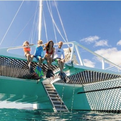 Four people sitting on the edge of a catamaran sailboat over clear blue water.