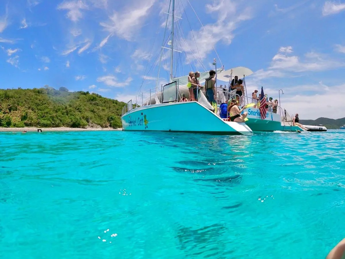 People on a turquoise boat in clear blue water near green hills.