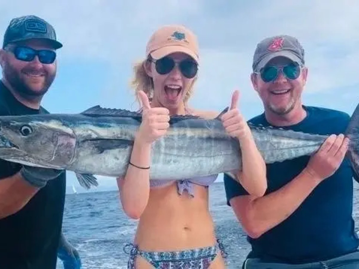 Three people holding a large fish on a boat, ocean in the background.