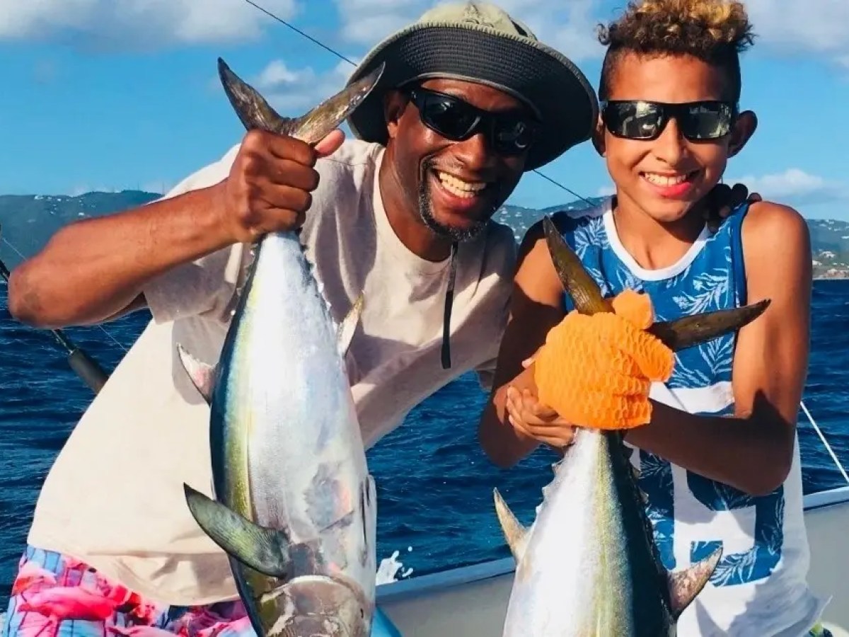 Two people smiling on a boat, holding large fish with the ocean in the background.