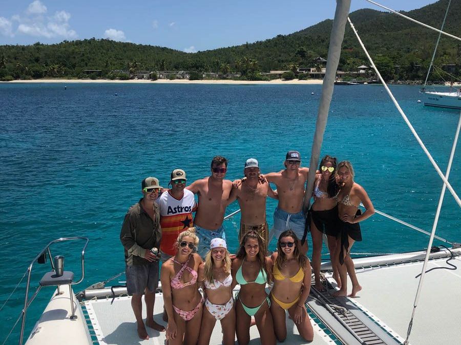 Group of people posing on a sailboat with clear blue water and an island in the background.