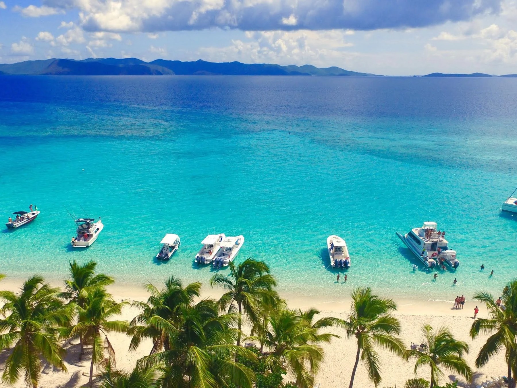 Tropical beach with calm turquoise water, boats, palm trees, and distant mountains.